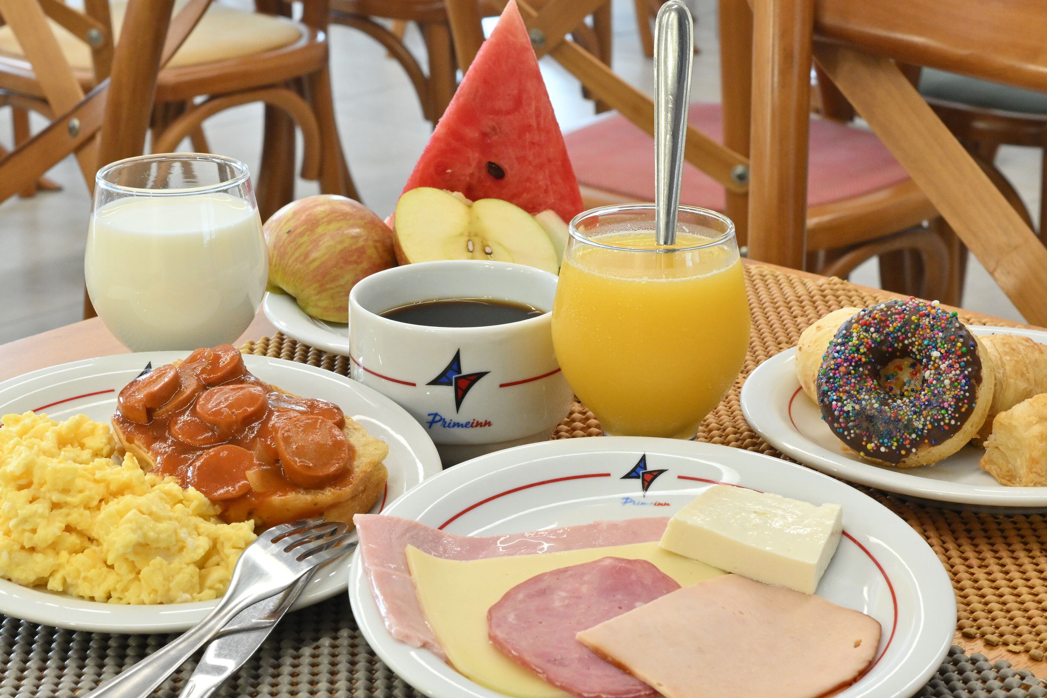 Une table de petit-déjeuner avec des assiettes d’œufs, de jambon, de fromage, de saucisses, des toasts et une tranche de pastèque. On y trouve également des verres de lait et de jus d’orange, ainsi qu’une tasse de café.