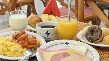 Une table de petit-déjeuner avec des assiettes d’œufs, de jambon, de fromage, de saucisses, des toasts et une tranche de pastèque. On y trouve également des verres de lait et de jus d’orange, ainsi qu’une tasse de café.