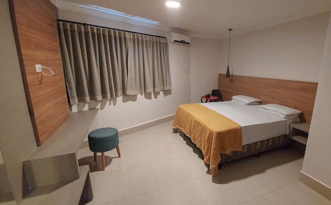 A hotel room with a bed, a small stool, a desk, and a window with curtains.
