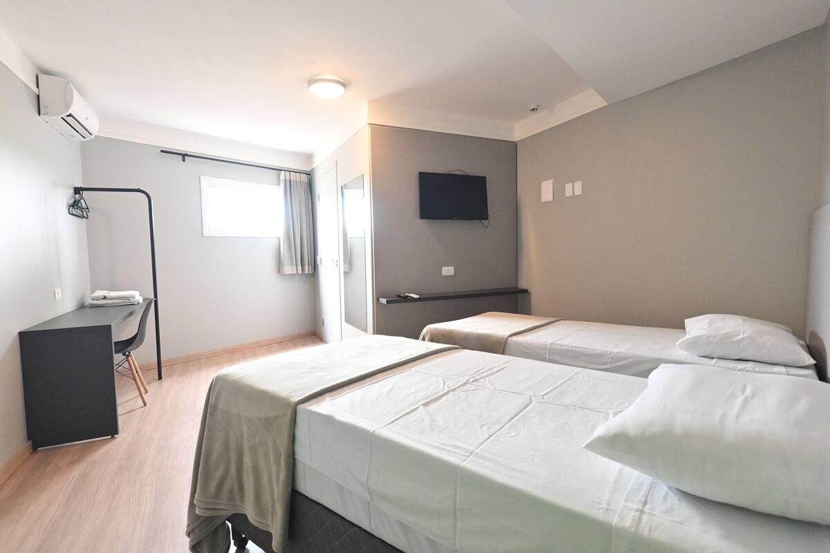 A hotel room with two beds, a desk, a chair, a TV, and a window with curtains.