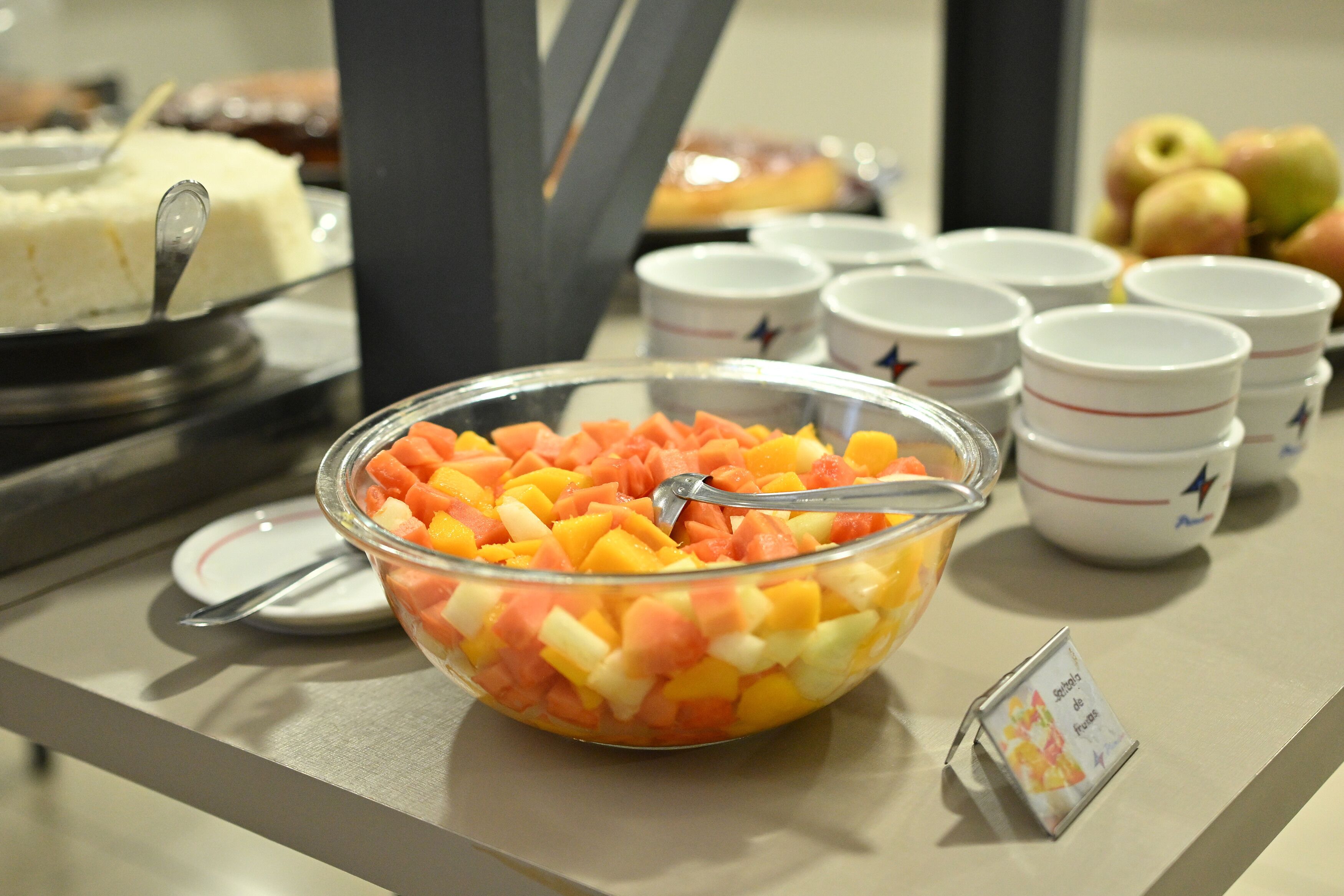 A buffet table with a bowl of mixed fruit, white cups with star designs, and a cake on a stand.