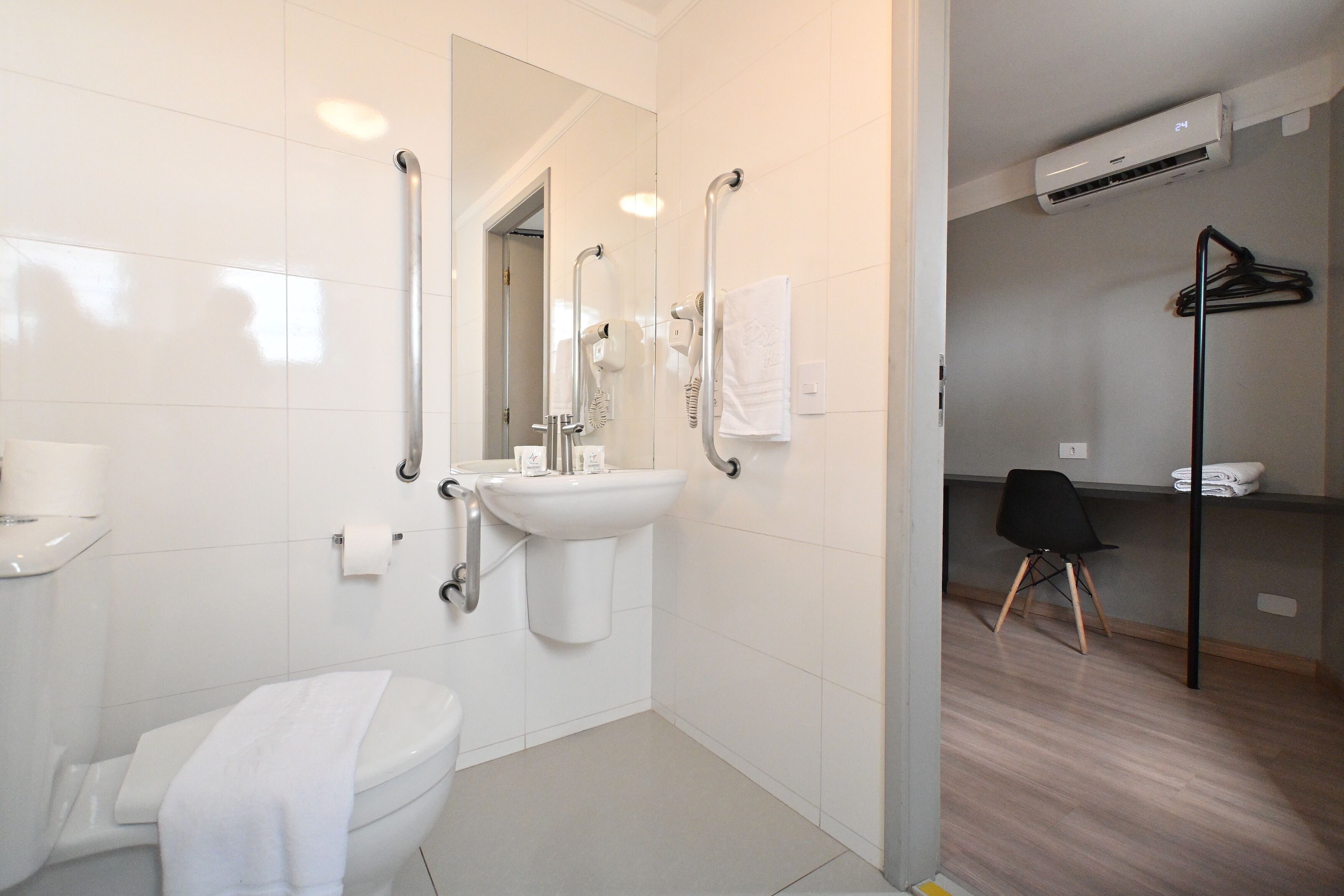 A modern bathroom with a toilet, sink, and mirror. There is a chair and a shelf with towels.