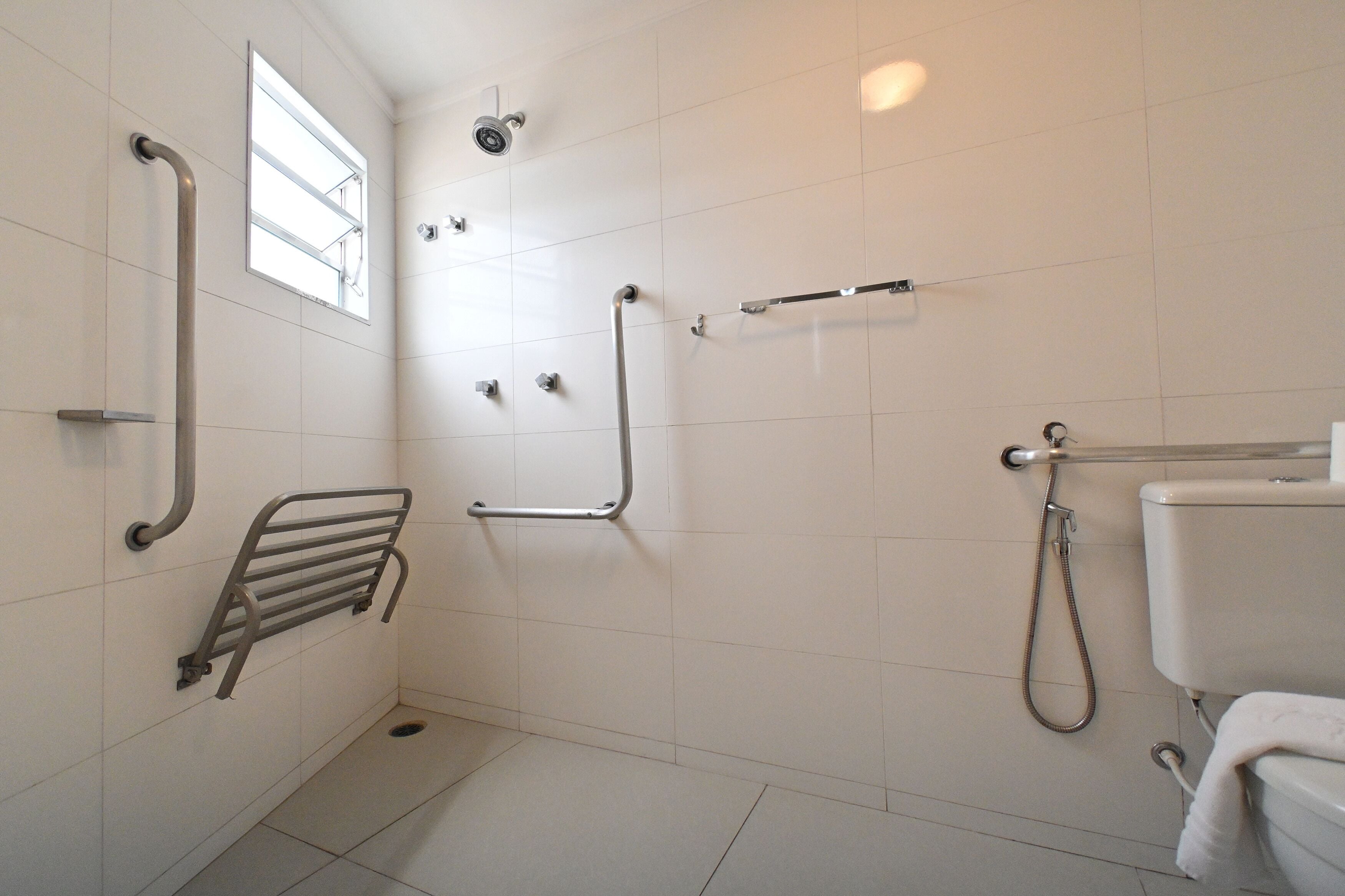 Un baño con azulejos blancos, inodoro, ducha con cabezal de ducha manual y barras de apoyo.