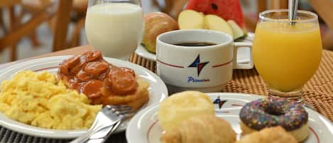 Un desayuno con platos de huevos revueltos, salchichas, croissants y un muffin, junto con un vaso de jugo de naranja y una taza de café.