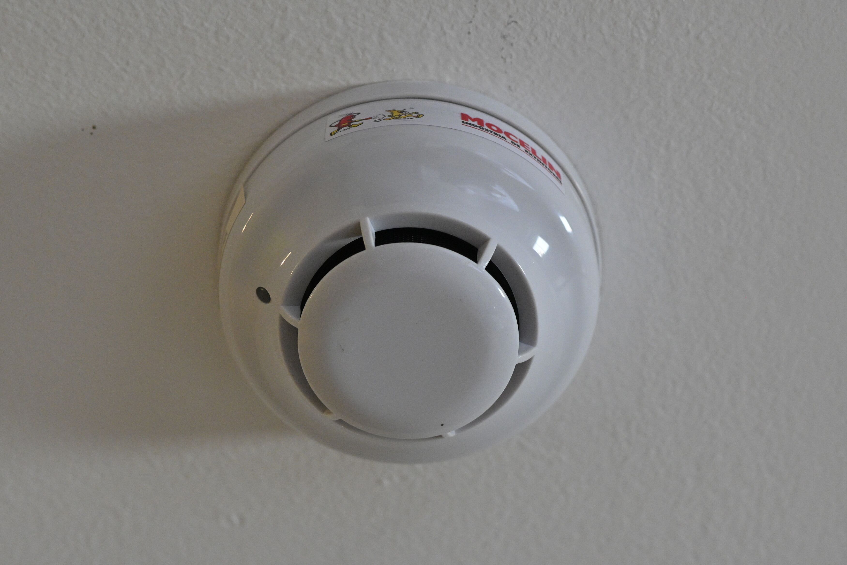 A white ceiling-mounted smoke detector with a round design and a sticker on the top.