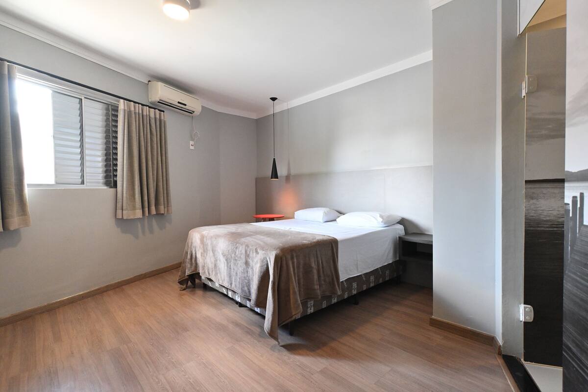 A modern hotel room with a bed, a bedside table, an air conditioning unit, a window with curtains, and a door.