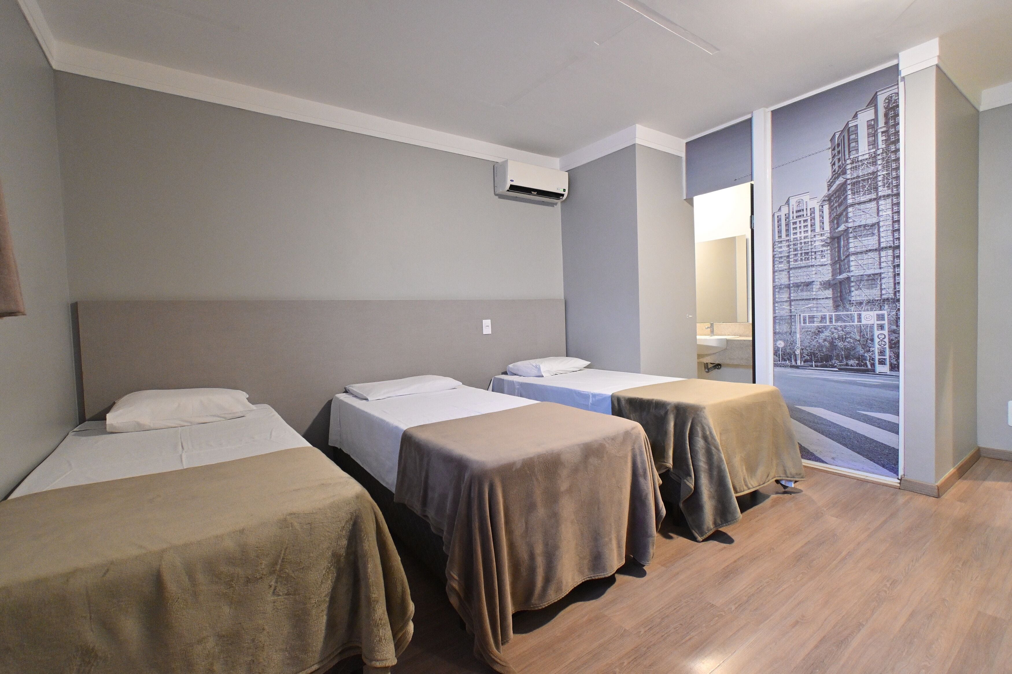 Habitació d'hotel amb tres llits, una finestra gran i vista a la ciutat, i un terra de fusta.