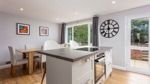 A modern kitchen with a central island, a dining area with chairs, a large window with curtains, and a wall-mounted clock. - Leeward House - Donnini Apartments (Ayr)