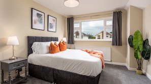 A bedroom with a bed, bedside table, lamp, window with curtains, radiator, and a plant.