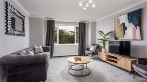 A modern living room with a grey sofa, a round coffee table, a TV stand, and a large window with curtains.