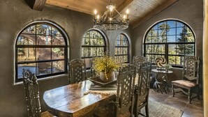 A dining area with a wooden table, ornate chairs, and a chandelier. Large arched windows provide views of the outdoors. - Unique mountain home with hot tub (Angel Fire)