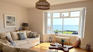 A living room with a beige sofa, a small wooden table, and a view of the sea through large windows.
