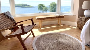 A room with a large window offering a view of a body of water and greenery. There is a wicker chair, a small bench, and a round rug on the wooden floor.