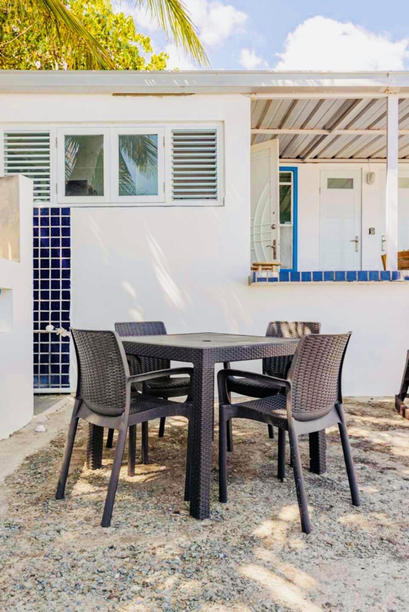 A covered outdoor dining area with a table and chairs, a white building with a blue door, and a palm tree in the background.