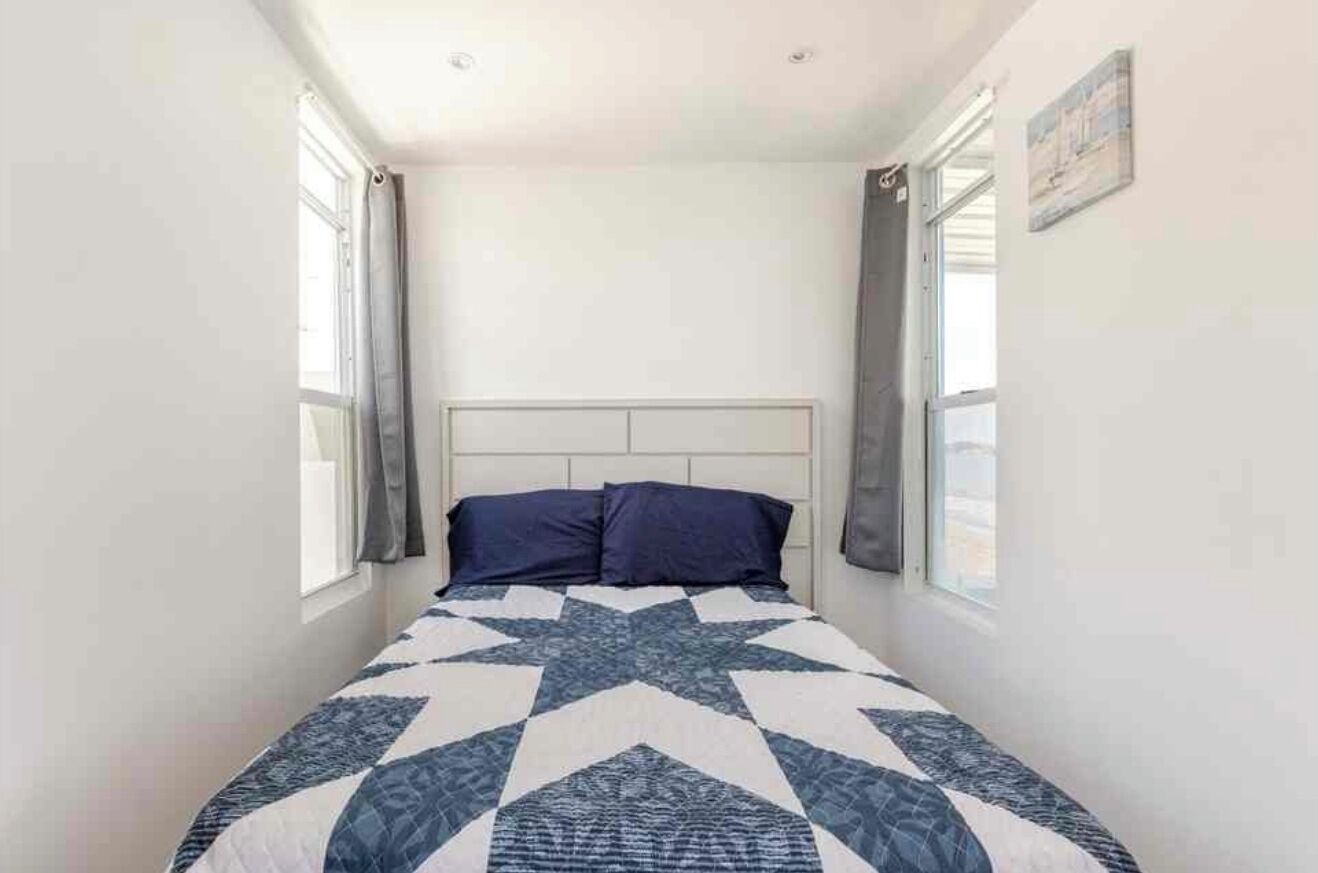 Een slaapkamer met een bed, hoofdeinde en twee ramen met gordijnen.