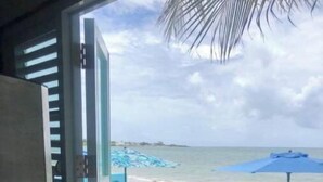 Une vue sur la plage, prise depuis une fenêtre, avec un parasol bleu et un palmier.