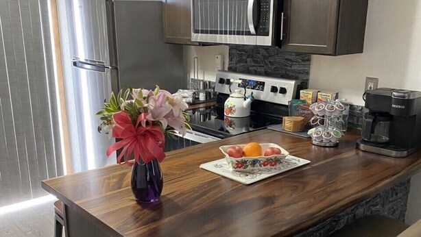 A modern kitchen with dark wooden cabinets, a stainless steel refrigerator, a microwave, a stove, and a coffee maker. There is a wooden island with a vase of flowers and a plate of fruit. - Elegant & Cozy 2BR Apartment with Chugach Mountain Views #1 (Anchorage)