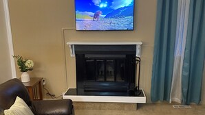 A living room with a fireplace, a television mounted above it, and a brown leather sofa. - Elegant & Cozy 2BR Apartment with Chugach Mountain Views #1 (Anchorage)