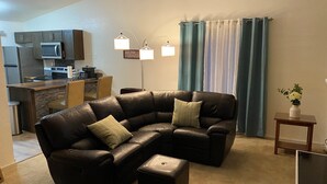 A living room with a sectional sofa, a small ottoman, a side table with a vase, and a kitchen area in the background. - Elegant & Cozy 2BR Apartment with Chugach Mountain Views #1 (Anchorage)