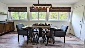 A dining area with a wooden table and chairs, large windows with blinds, and a kitchen area with cabinets and a countertop. - An Updated, Secluded family friendly home with Fast Wifi (Manhattan)
