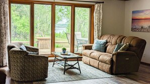 A living room with a brown sofa, patterned armchair, and a coffee table. Large windows offer a view of greenery outside. - An Updated, Secluded family friendly home with Fast Wifi (Manhattan)