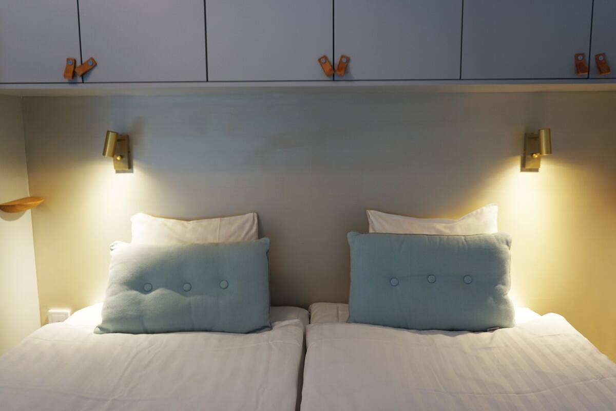 A double bed with two pillows, a nightstand, and a wall-mounted shelf with two orange tags.