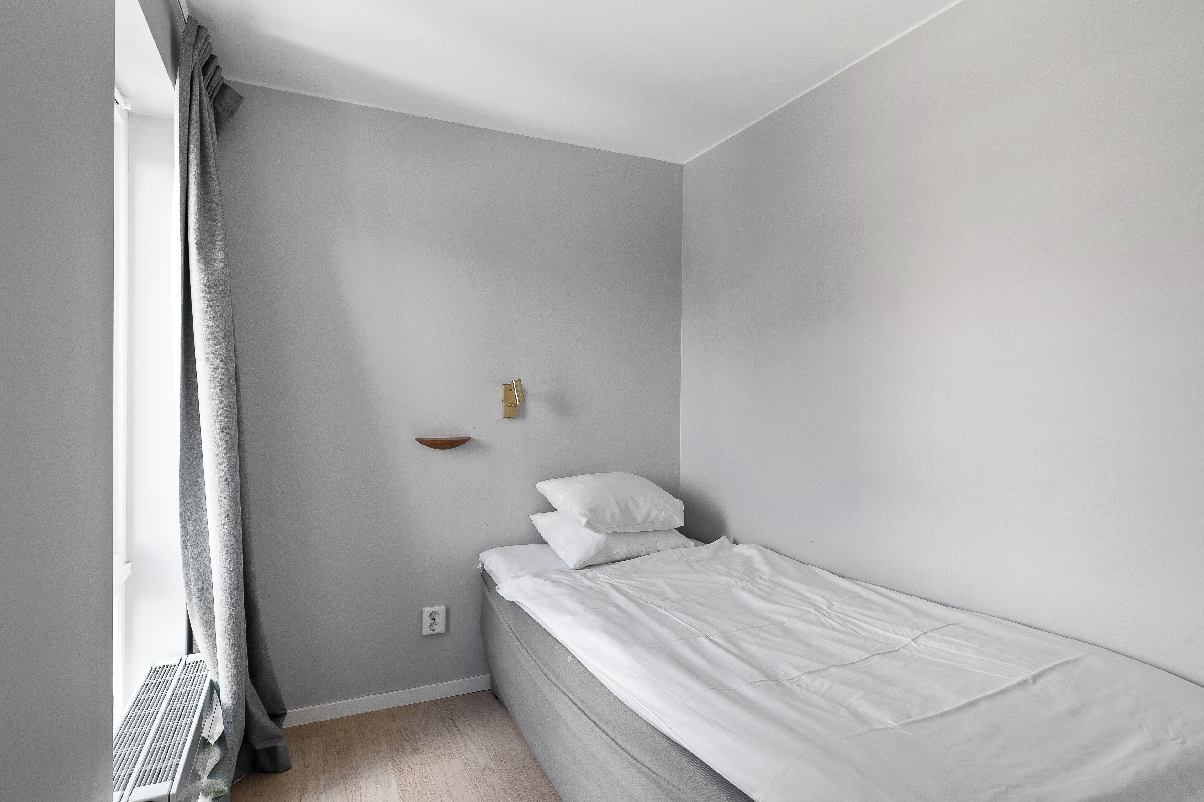 A small, neatly made bed in a room with light gray walls, a radiator, and a small shelf with a light fixture.