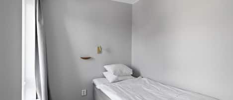 A small, neatly made bed in a room with light gray walls, a radiator, and a small shelf with a light fixture.