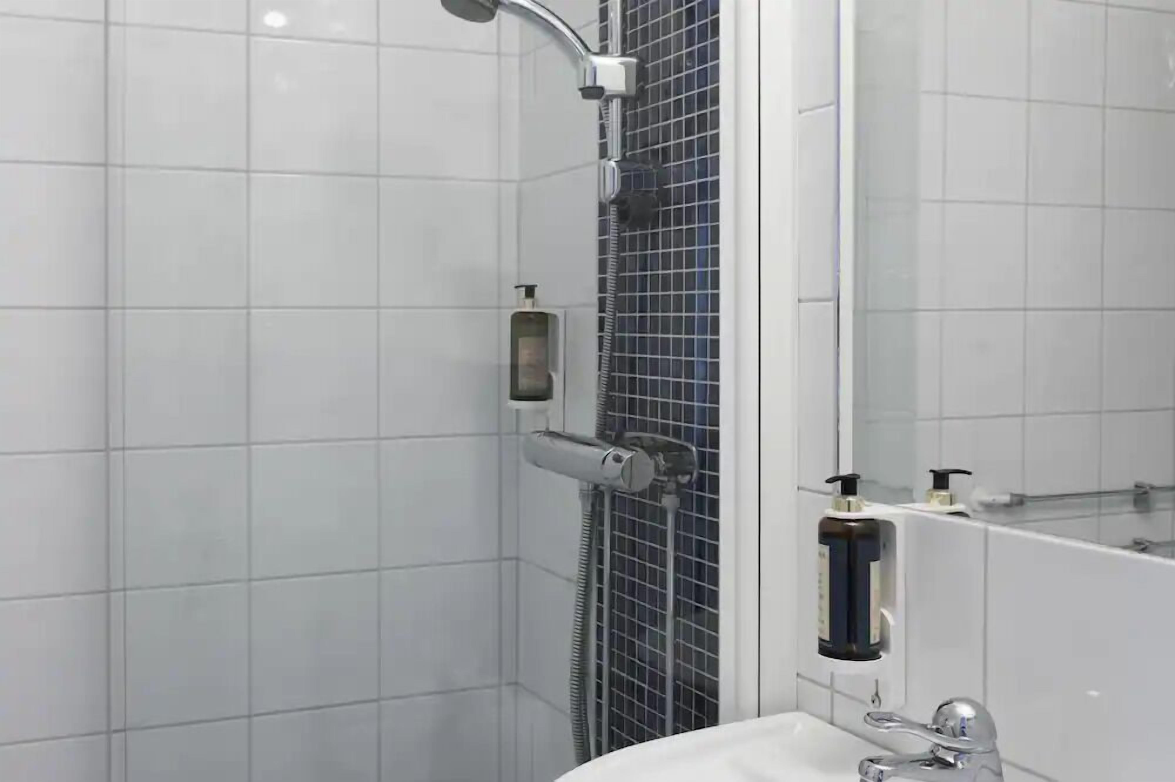 A modern bathroom with white tiles, a shower with a handheld showerhead, a glass-enclosed shower, and a mirror above the sink.