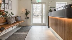 A reception area with a wooden reception desk, a bench with cushions, a potted plant, and framed pictures on the wall. - Avanti Hotel (Hägersten)
