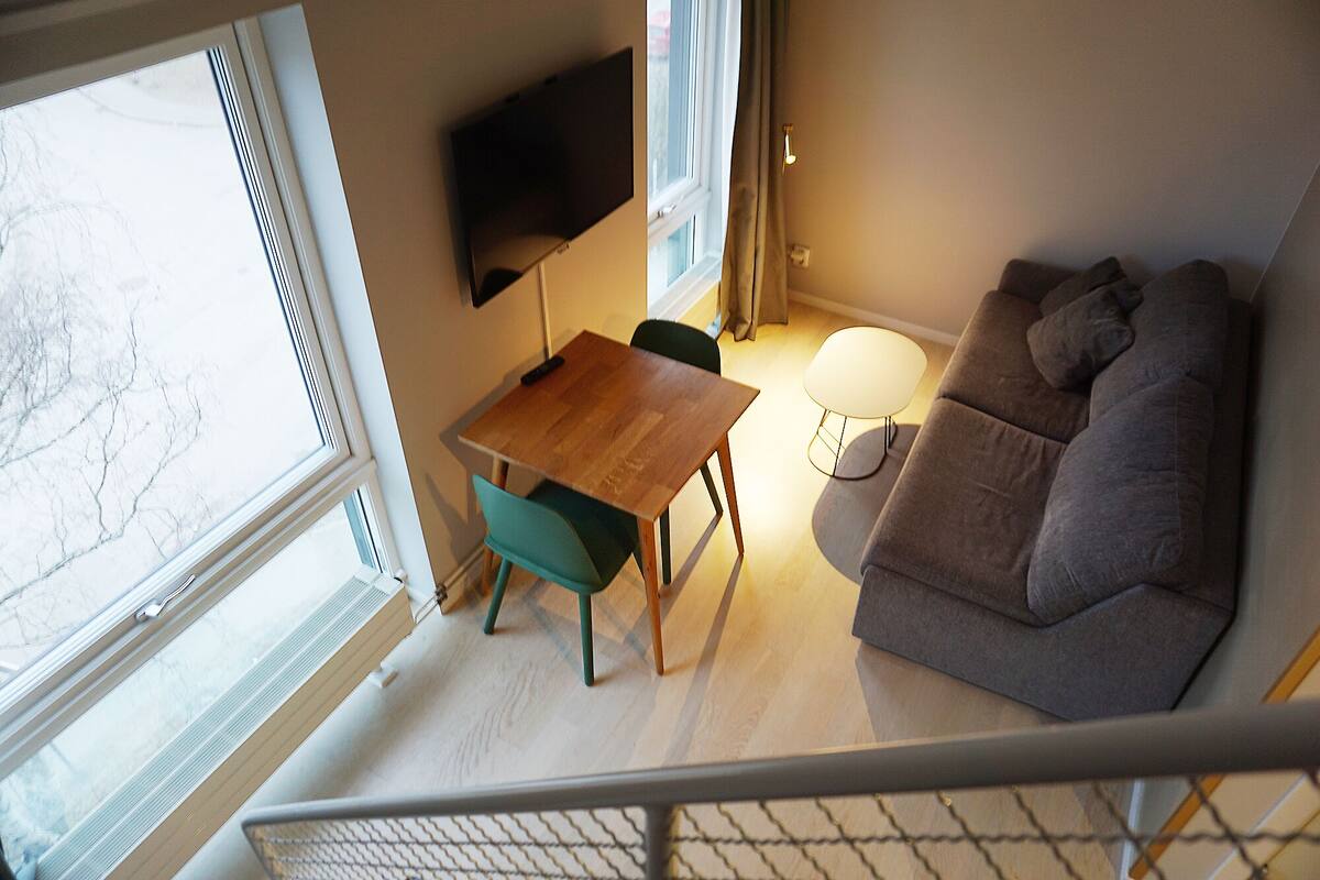 A living room with a sofa, a dining table with chairs, a TV mounted on the wall, and a window.