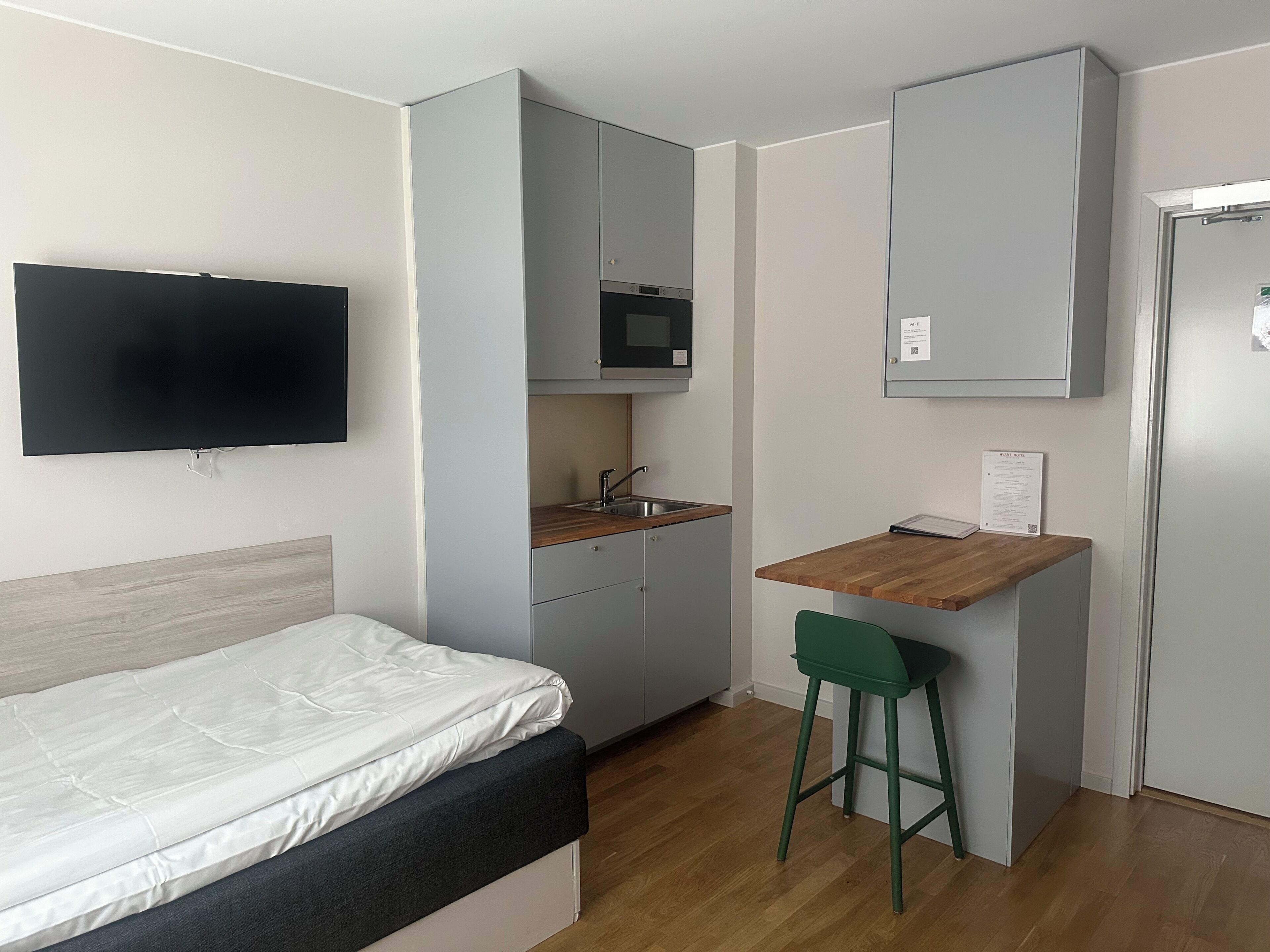 A compact room with a bed, a kitchenette, a small dining area, and a TV mounted on the wall.