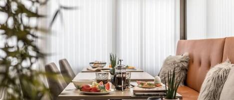A modern dining area with a wooden table set for two, featuring a fruit platter, a coffee pot, and a glass pitcher.