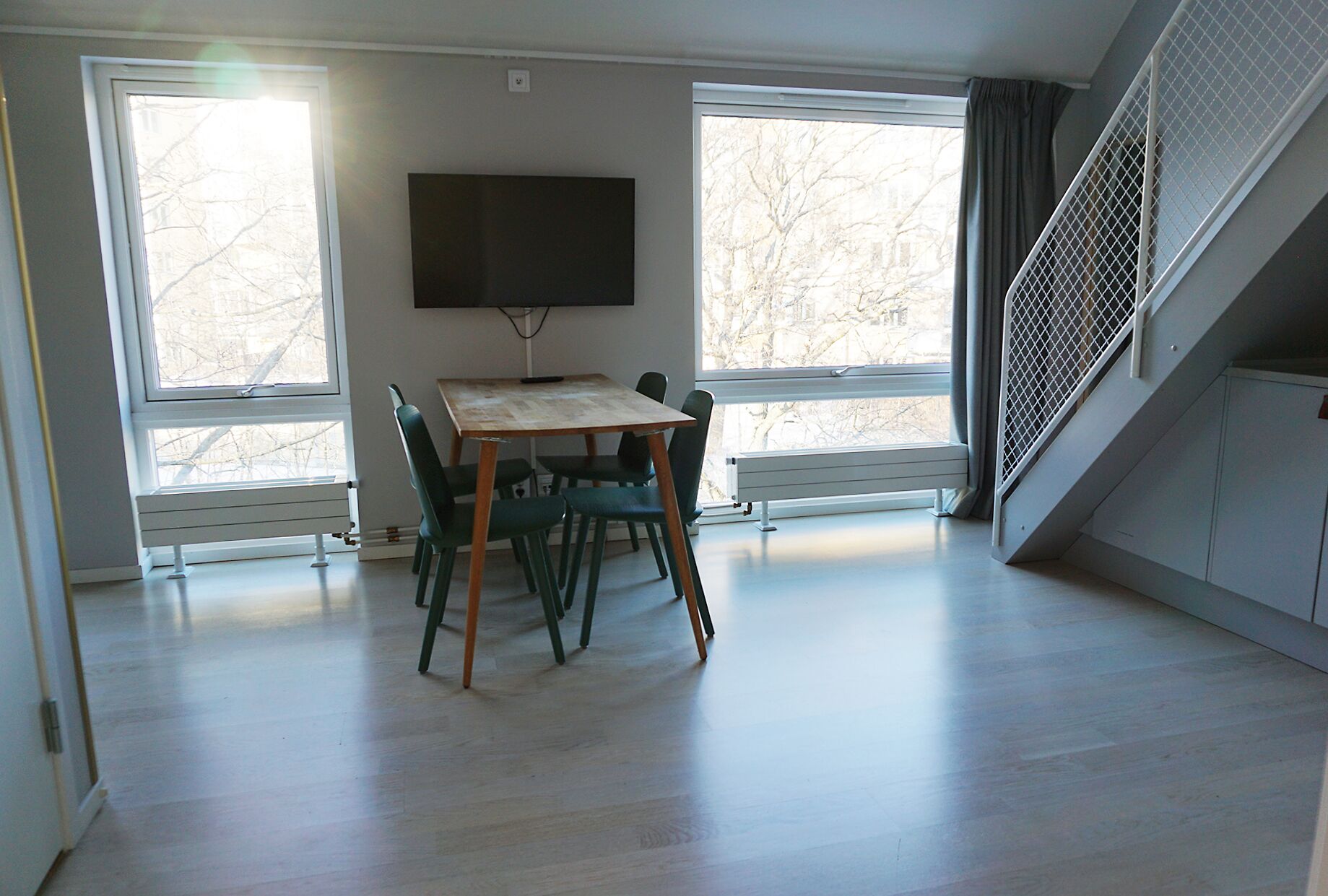 Una habitación con una mesa y sillas de madera, un televisor de pantalla plana en la pared y ventanas amplias con vistas a árboles desnudos.