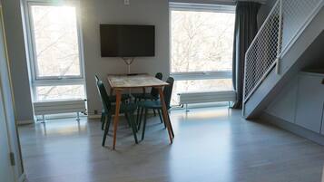 Una habitación con una mesa y sillas de madera, un televisor de pantalla plana en la pared y ventanas amplias con vistas a árboles desnudos.