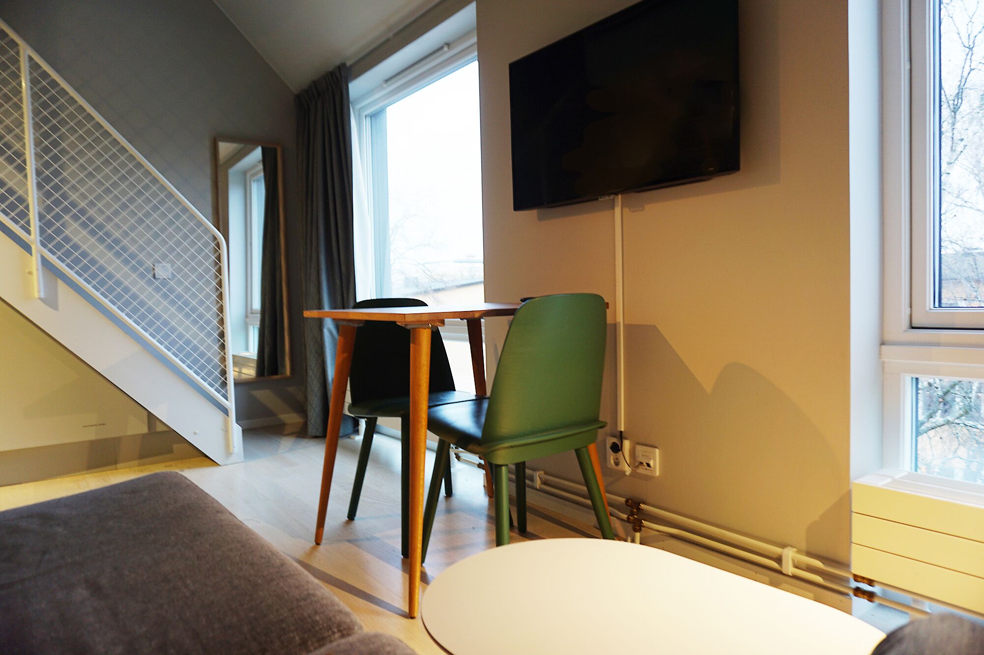 A hotel room with a staircase, a TV mounted on the wall, a dining table with chairs, and a window with a view.