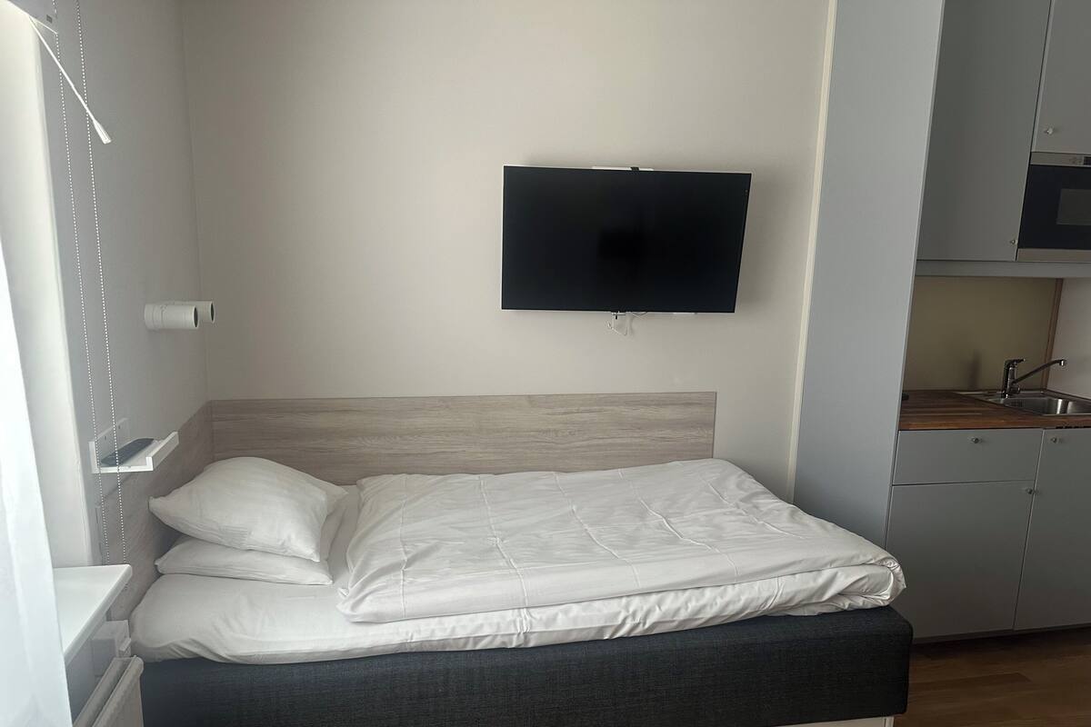 A bedroom with a bed, a TV mounted on the wall, a wooden headboard, and a kitchenette area.