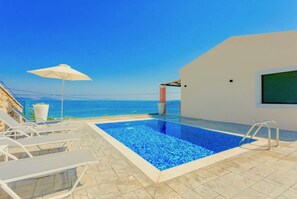 A swimming pool with lounge chairs, a white umbrella, and a view of the sea.