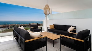 Ein Balkon mit einer schwarzen Couch, zwei schwarzen Sesseln, einem Holztisch und einer hängenden Lampe.
