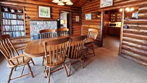 A rustic wooden interior with a dining area featuring a large wooden table and chairs, a stone fireplace, and a bookshelf filled with books. - Lakeside Lodge on Lake Huron (Saint Ignace)