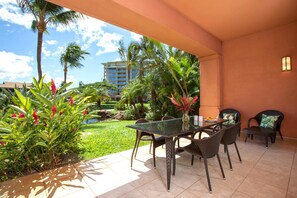 Um pátio externo com mesa de vidro, cadeiras de vime e um pequeno jardim com plantas tropicais e um recurso de água.