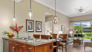 A modern kitchen with granite countertops, a dining area with a wooden table and chairs, and a living room with a sofa and a view of a tropical landscape. - Honua Kai | Ocean View 2 BR Sleeps 6 | Car Incl. w/ 6+ Nights | HKK-112 by KBM (Lahaina)