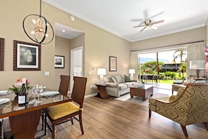 A spacious living area with a dining table set for two, a sofa, and a coffee table. There is a ceiling fan, a large window with a view of the outdoors, and a door leading to another room. - Honua Kai | Mountain View 2 BR | Free Car w/ 6+ Nights | HKK-112 (Lahaina)
