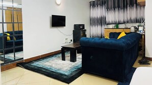 A living room with a blue sofa, a flat-screen TV, a small table, and a kitchen area with a microwave. - Beautiful & cosy studio, 5 mins from two rivers (Ruaka)