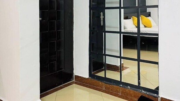 A room with a sliding glass door leading to a bed with white bedding and yellow pillows. The room has a tiled floor and a blue rug. - Beautiful & cosy studio, 5 mins from two rivers (Ruaka)