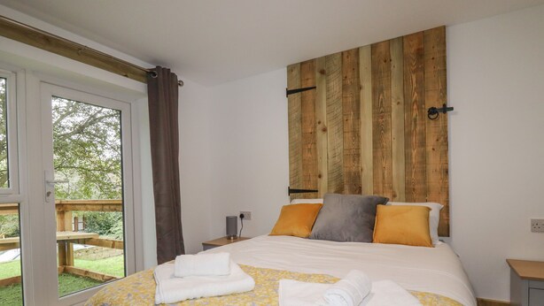 A bedroom with a wooden headboard, a bed with a yellow floral bedspread, two pillows, and a set of towels. - Stall 2 - The Stables (Truro)