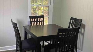 A dining area with a rectangular table and four chairs, a window with a view of greenery, and wooden flooring. - Camden Cottage (Camden)