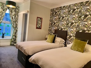 Habitación de hotel con dos camas, un ventanal con cortinas, una pared con estampado de hojas tropicales y un cuadro pequeño en la pared.