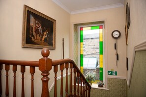 Een trap met houten leuning, een ingelijst schilderij aan de muur, een raam met uitzicht op strepen in groen en geel, en een klok aan de wand.