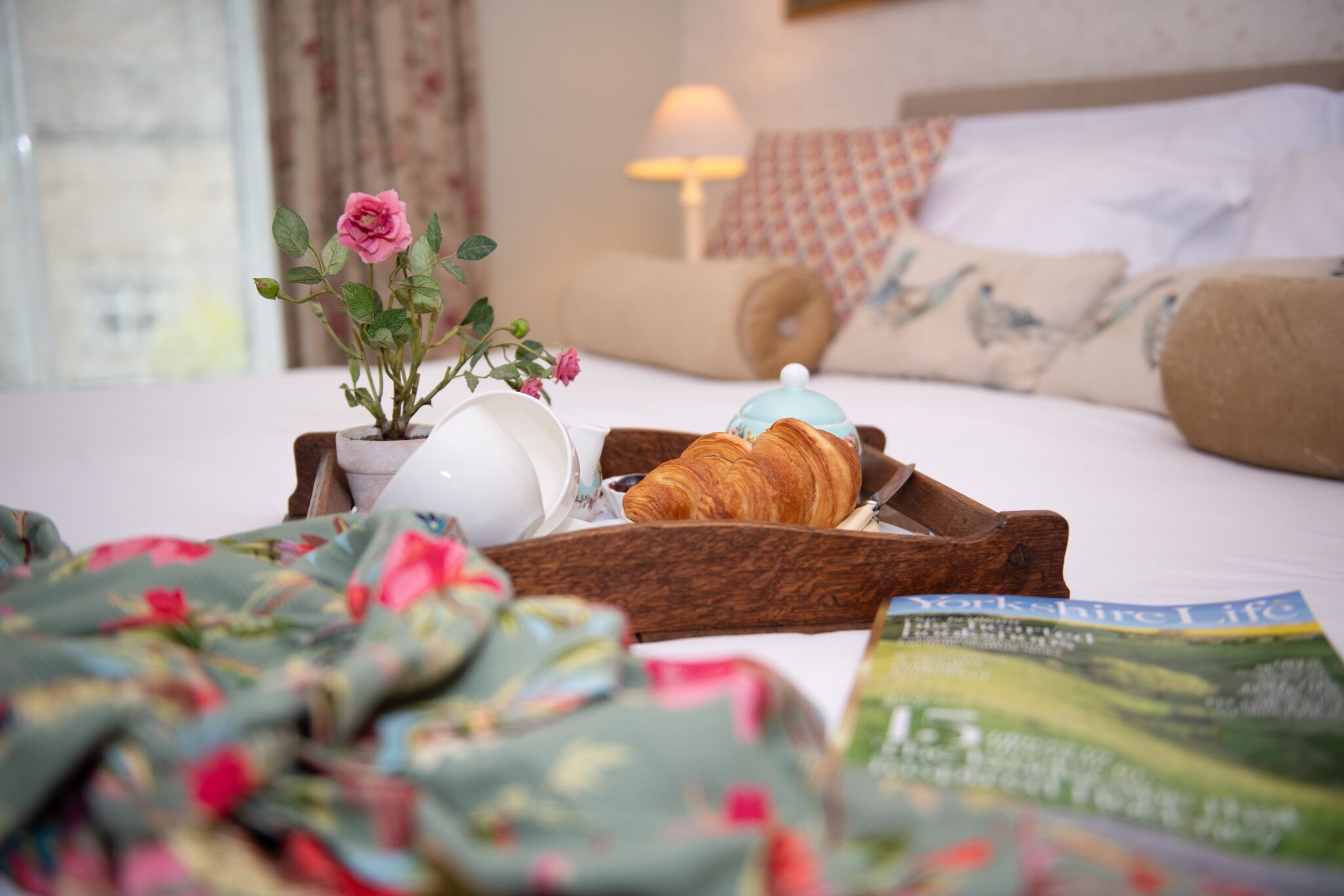 Ein ordentlich bezogenes Bett mit Kissen, ein Nachttisch mit Lampe, ein Tablett mit Croissant, Tasse und einer kleinen Topfpflanze.